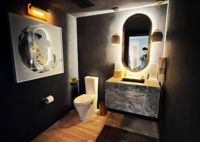 A modern bathroom with dark walls, featuring a toilet, an illuminated mirror above a marble sink, hanging pendant lights, a contemporary piece of wall art, and a wooden floor.