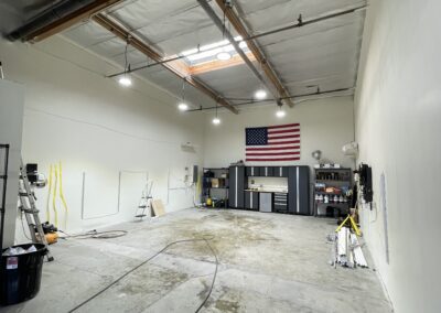 A spacious garage with an American flag on the wall, industrial lighting, shelves with tools, and vehicles partially visible. The floor appears wet with a hose and various equipment scattered around.