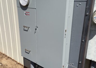 An outdoor electrical meter box installed on a wall next to a grey pipe, with labeled compartments and a round meter. The surrounding ground is brown dirt.