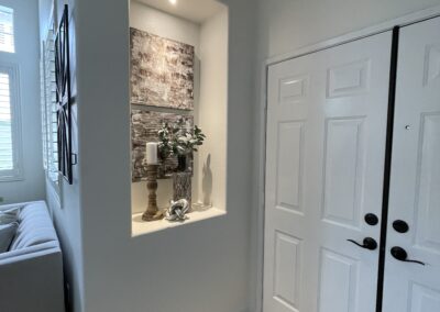 A small recessed alcove in an entryway features decorative items including a candle holder, plants, and artwork. The alcove is illuminated by a small light, and a double door is visible to the right.