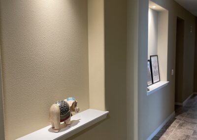 A hallway with beige walls, recessed lighting, and tiled flooring. A small decorative elephant figurine is placed on a built-in shelf on the left, and framed art stands on a built-in shelf farther down the hall.