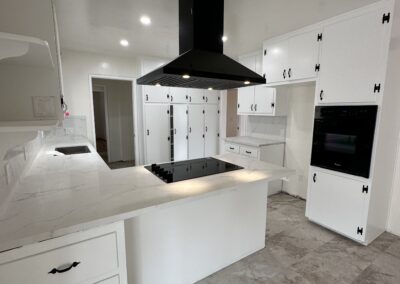 A modern, white kitchen with a central island, black range hood, and built-in oven. Marble countertops and tiled floor are present. Cabinets and drawers line the walls.