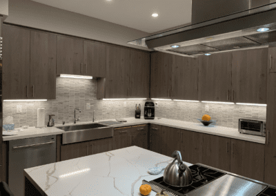 Modern kitchen with stainless steel appliances, dark wood cabinets, and a marble countertop. Backsplash is made of white tiles. A single orange and a kettle are on the countertop.