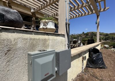 Outdoor electrical panels attached to the side of a building with a pergola above, various garden items, and a scenic landscape in the background.