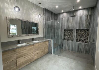 Modern bathroom with a large walk-in shower featuring gray marble walls and two accent niches. A double sink vanity with wooden cabinets is on the left, and pendant lights hang above a large mirror.