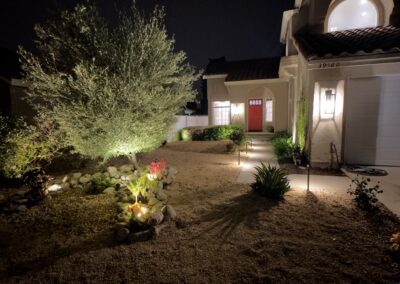 A two-story house with lit exterior at night, featuring a red front door, illuminated tree and landscaped yard.