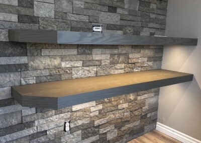 Two wall-mounted gray shelves on a stone wall. One shelf has electrical outlets above it, and the lower shelf has an electrical outlet directly below. The floor is wooden.